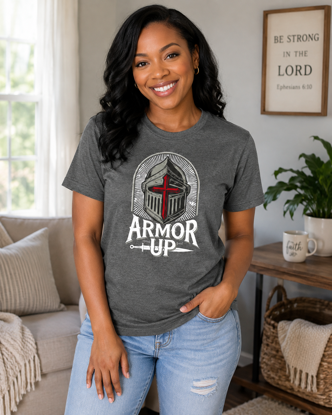 A woman wearing a dark heather gray graphic t-shirt with a knight helmet and the words 'ARMOR UP' printed on the front, paired with light blue jeans in a cozy living room setting.