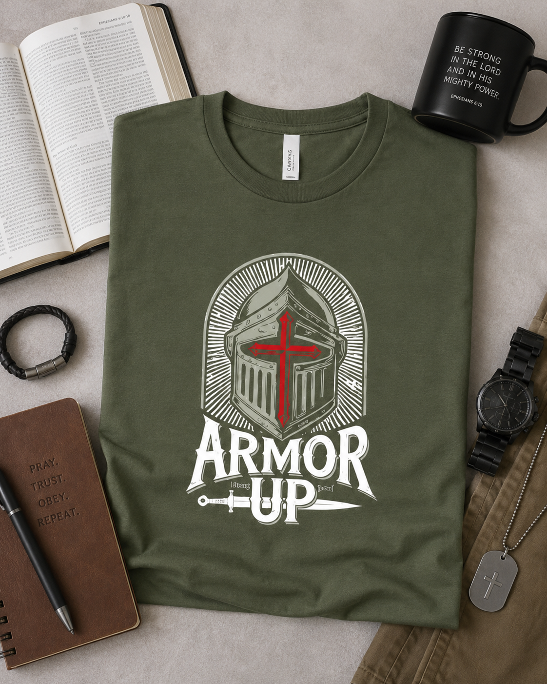An olive green graphic t-shirt featuring a white knight helmet with a red cross and the text 'ARMOR UP' underneath, styled in a flat-lay arrangement with a notebook, watch, mug, and other accessories on a neutral background.