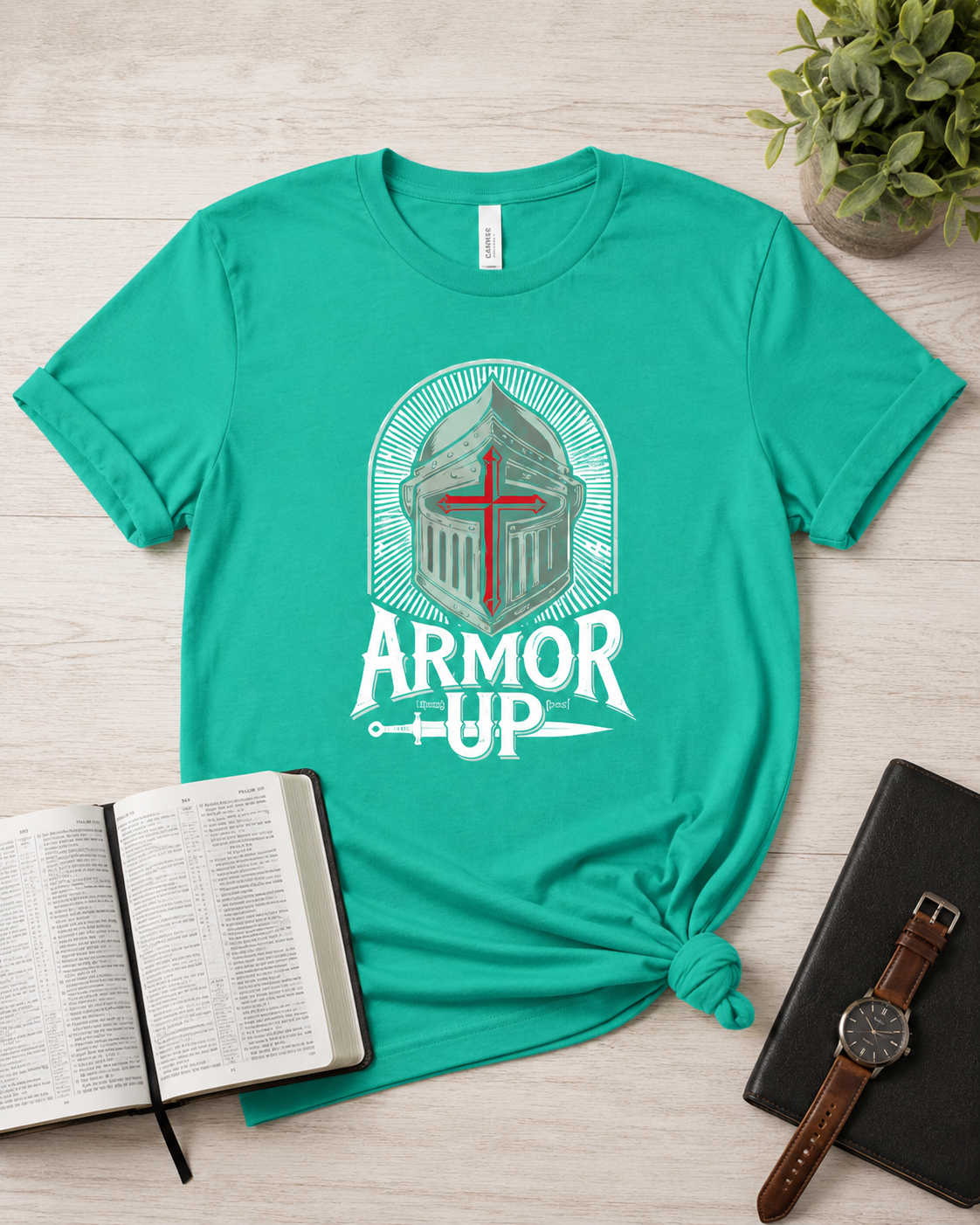 A teal short-sleeve graphic T-shirt laid flat on a light wood surface, styled with an open Bible, a black notebook with a brown leather watch, and a small potted plant. The shirt has a large front graphic showing a white and gray knight helmet with a red cross and the words “ARMOR UP.