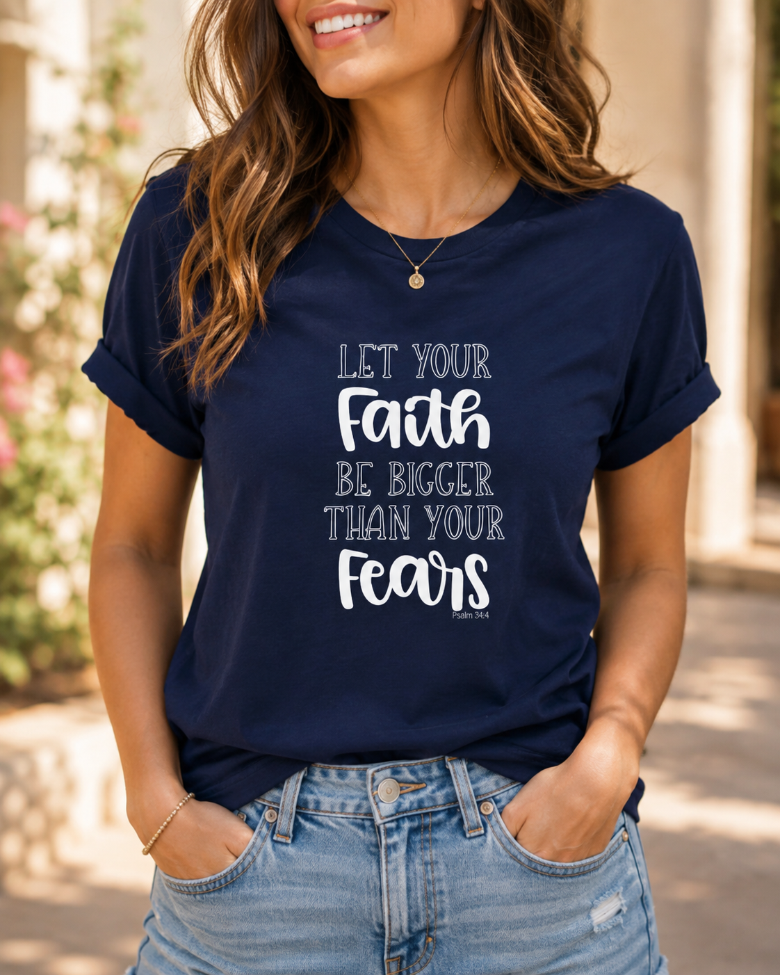 A woman wearing a navy blue graphic t-shirt with the text 'LET YOUR Faith BE BIGGER THAN YOUR fears' printed in white on the front, paired with light wash blue jeans.