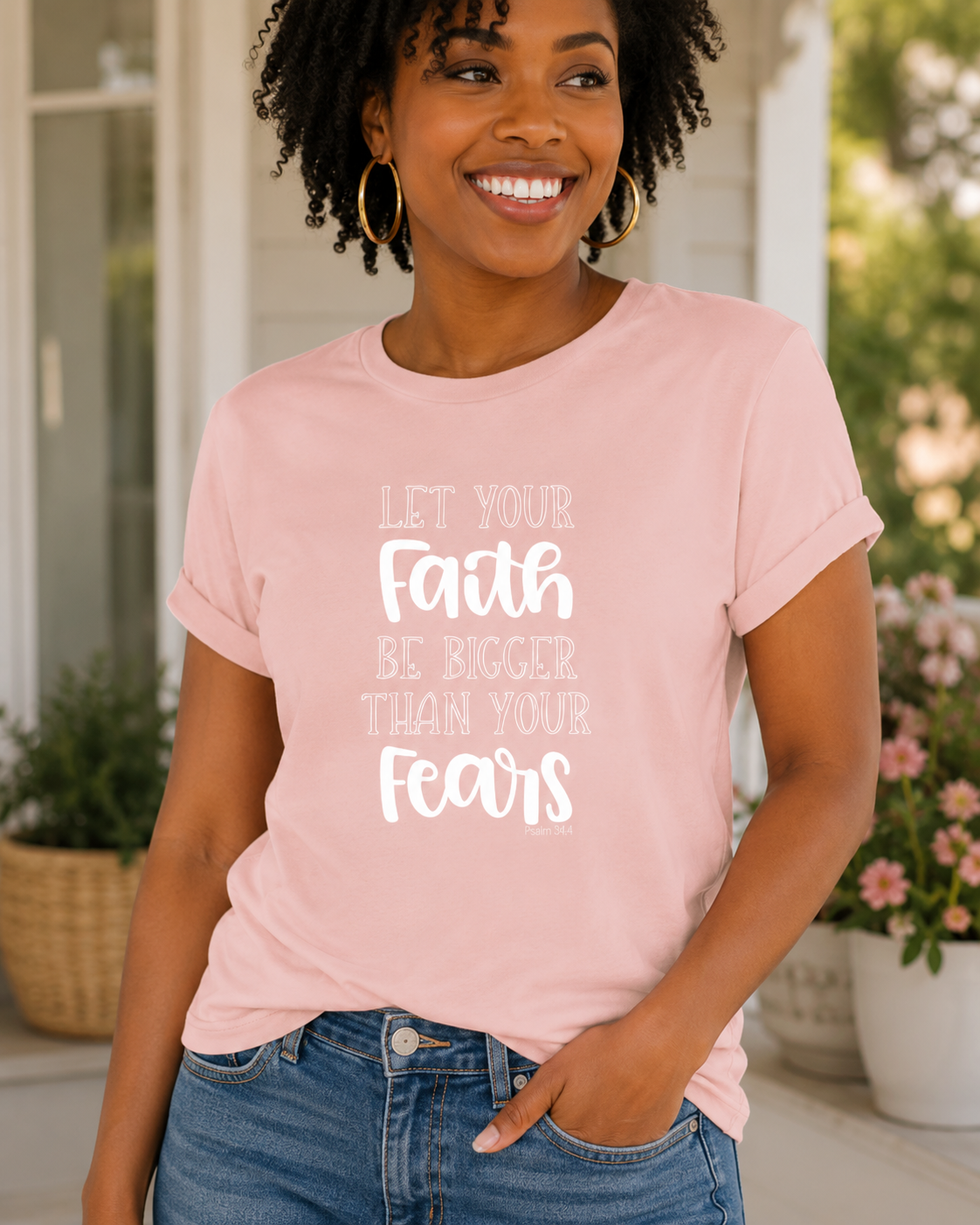 A smiling woman with curly hair wearing a light pink graphic t-shirt and blue jeans, standing on a porch with plants in the background. The t-shirt features the white text 'LET YOUR Faith BE BIGGER THAN YOUR Fears'.