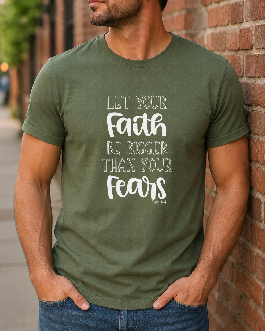 A man wearing an olive green t-shirt with a white printed quote that reads 'LET YOUR Faith BE BIGGER THAN YOUR Fears Phil. 4:13' standing in front of a brick wall.
