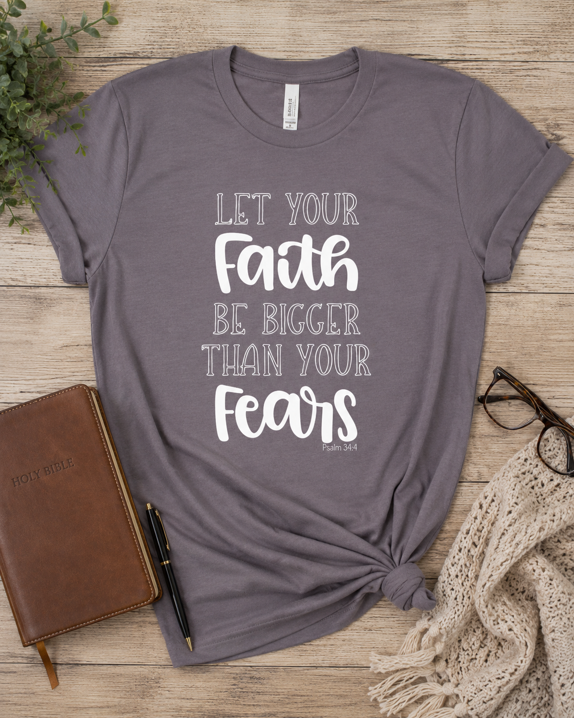 A heathered gray short-sleeve t-shirt with a white typographic print reading 'LET YOUR Faith BE BIGGER THAN YOUR Fears' and 'PSALM 37:5' at the bottom. The shirt is styled in a flat-lay on a light wood background with a brown leather journal, a black pen, eyeglasses, and a cream knit scarf.