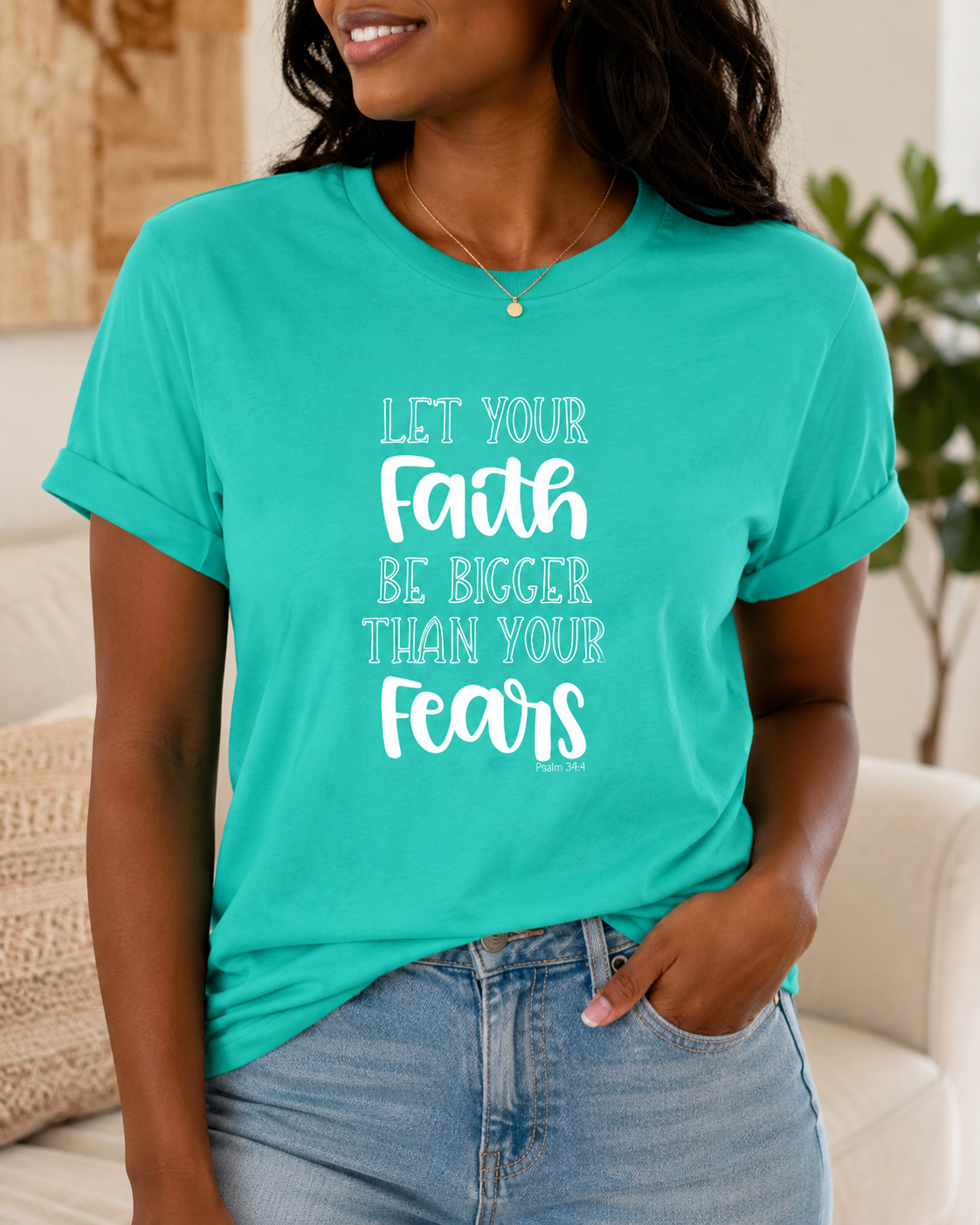 A woman wearing a teal t-shirt with the white printed text 'LET YOUR Faith BE BIGGER THAN YOUR Fears' and light blue jeans, shown in a cozy indoor setting.