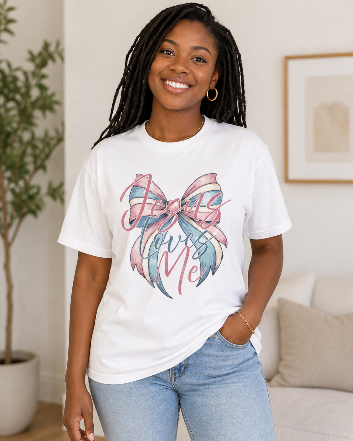 A smiling woman with long black dreadlocks models a white graphic t-shirt featuring a pink and blue bow design with the words 'Jesus Loves Me' in script, paired with light wash blue jeans in a bright, modern living room setting.