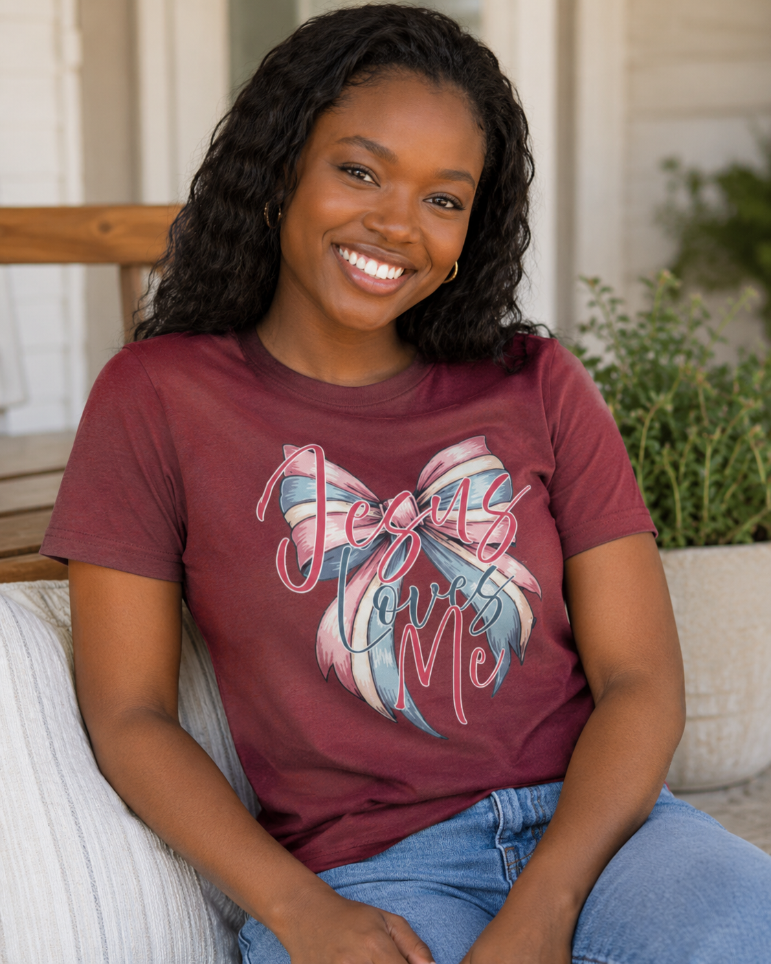 A smiling woman with long black curly hair sits on a porch wearing a maroon short-sleeve graphic T-shirt with a pastel ribbon and the words 'Jesus Loves Me' in pink script, paired with blue jeans.