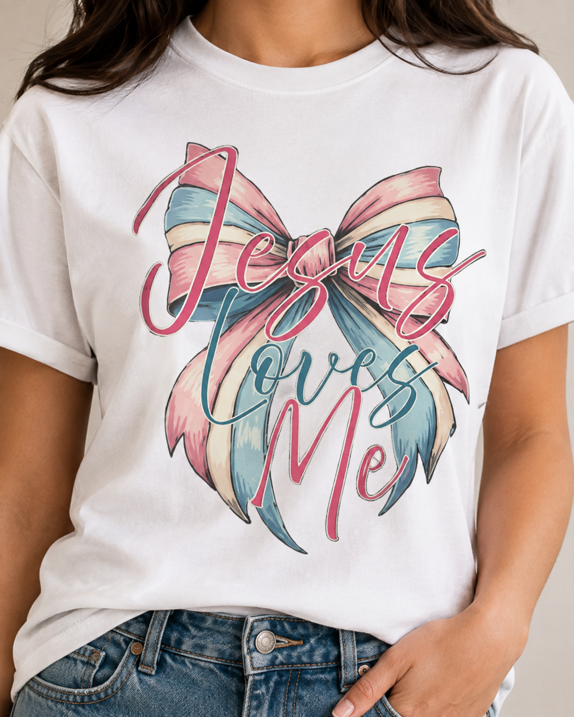 A white short-sleeve graphic t-shirt worn by a model, featuring a large bow illustration in pink, blue, and cream with the text 'Jesus Loves Me' written across it.