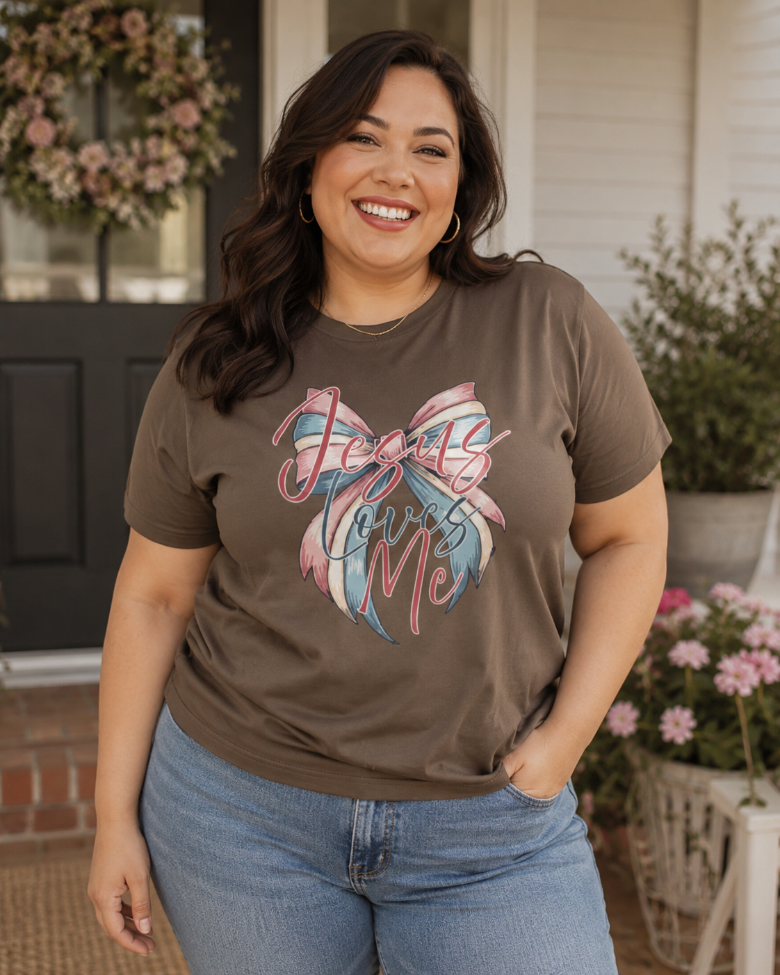 A smiling woman wearing a brown graphic t-shirt with a pink and blue ribbon heart design and the text 'Jesus Loves Me', paired with blue jeans, standing on a front porch.