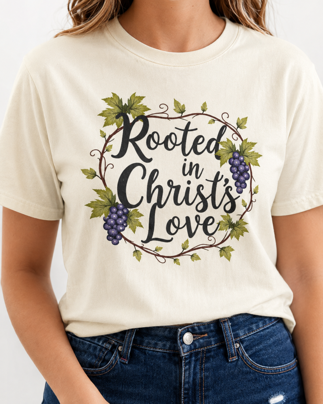 A woman wearing a cream-colored t-shirt with a graphic that reads 'Rooted in Christ's Love' in black script, surrounded by green leaves and purple grapes, paired with blue jeans.