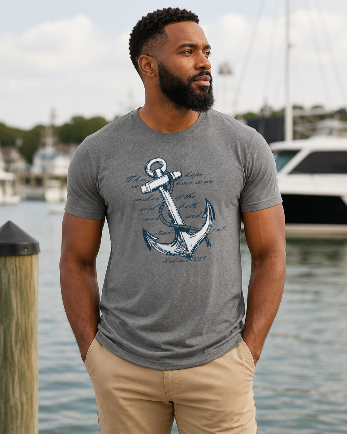 A man wearing a heather gray crewneck t-shirt with a blue anchor and script graphic, standing by a waterfront with boats in the background.