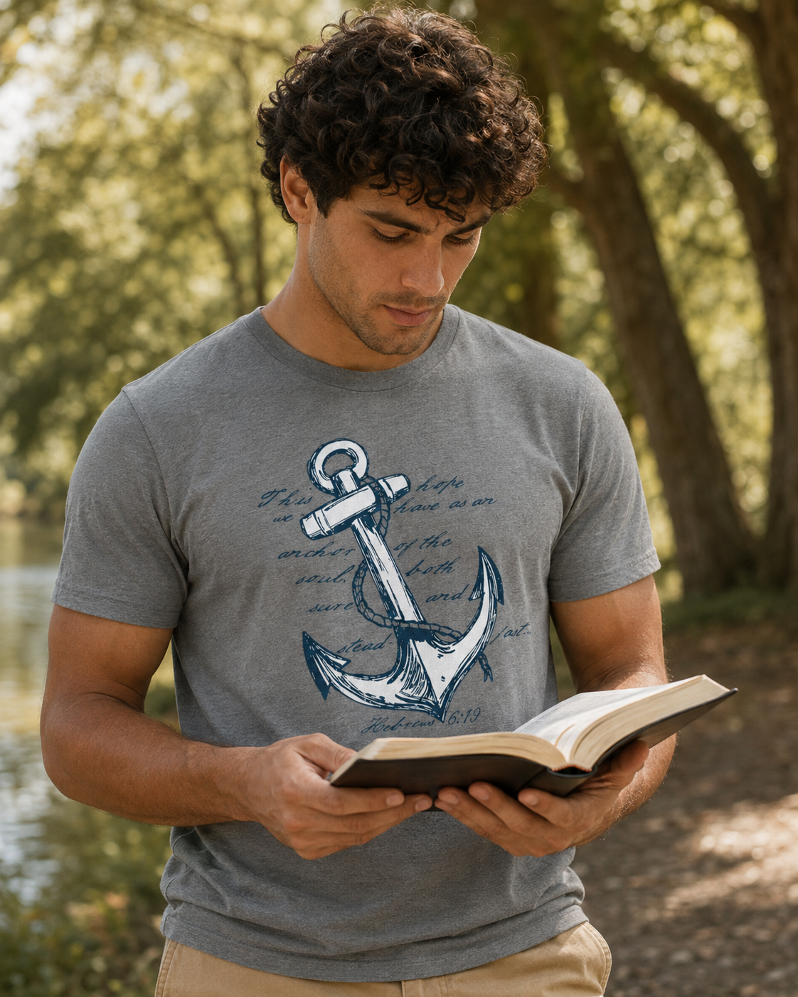 A man wearing a gray graphic t-shirt with a blue anchor design, standing outdoors in a wooded area and reading an open book.
