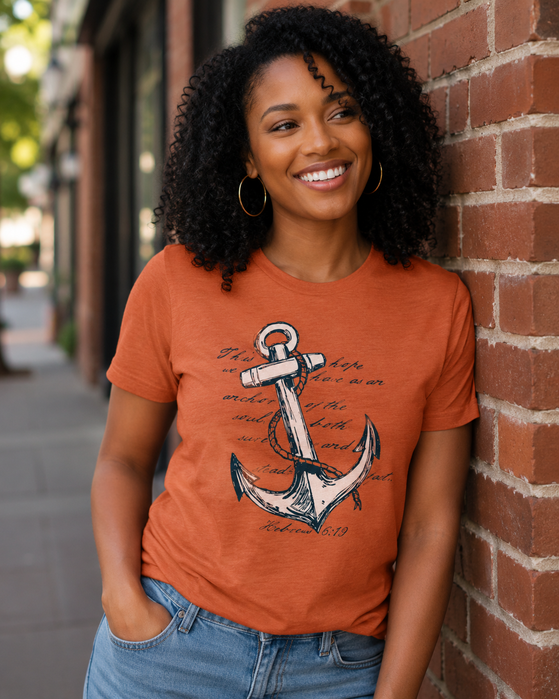 A woman with curly black hair smiling and leaning against a red brick wall, wearing a burnt orange graphic t-shirt with a white and navy anchor design, and blue jeans.