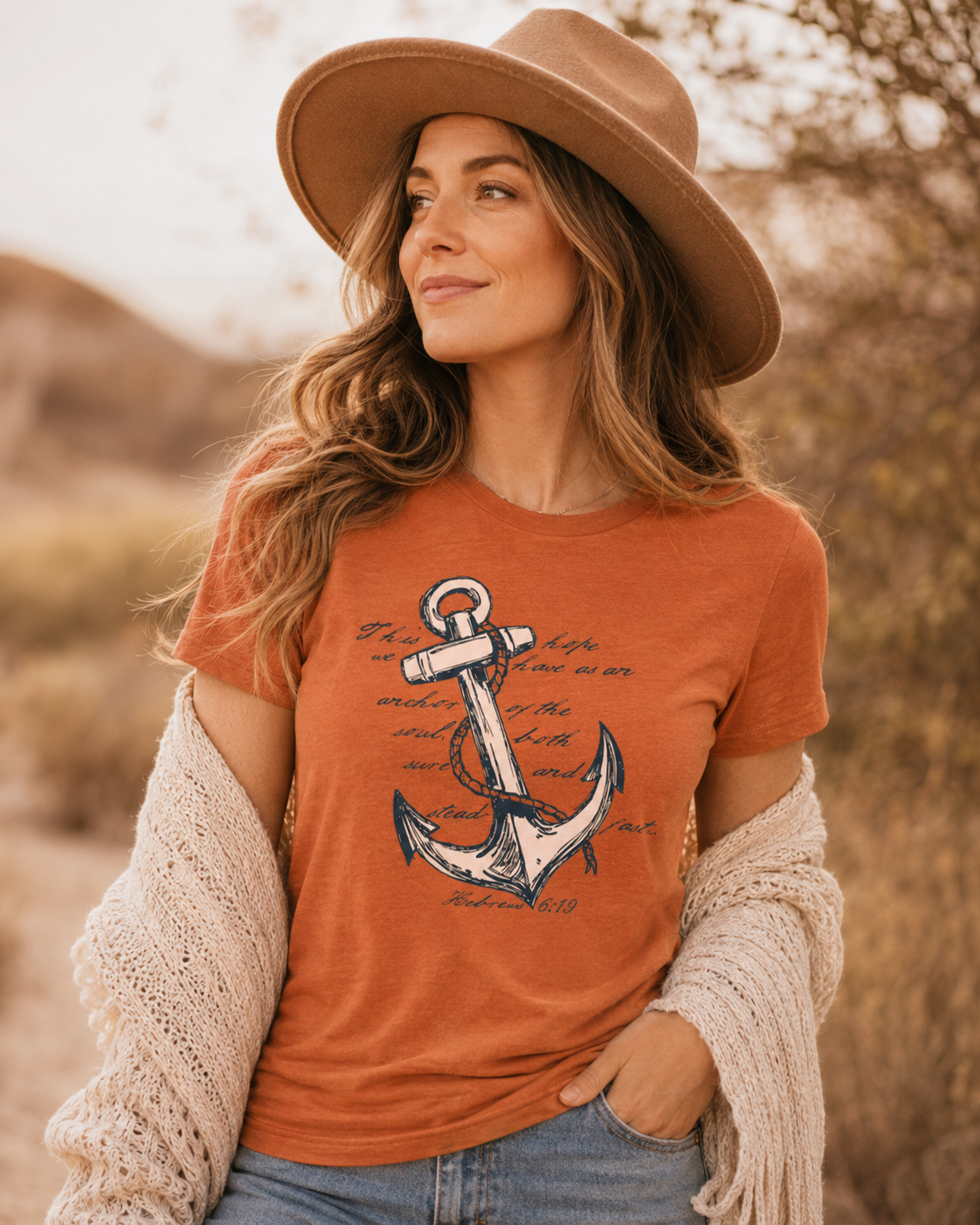 A woman wearing a rust-orange graphic t-shirt with a navy anchor print, a beige wide-brim hat, and a cream knit cardigan, posing outdoors in a warm desert landscape.