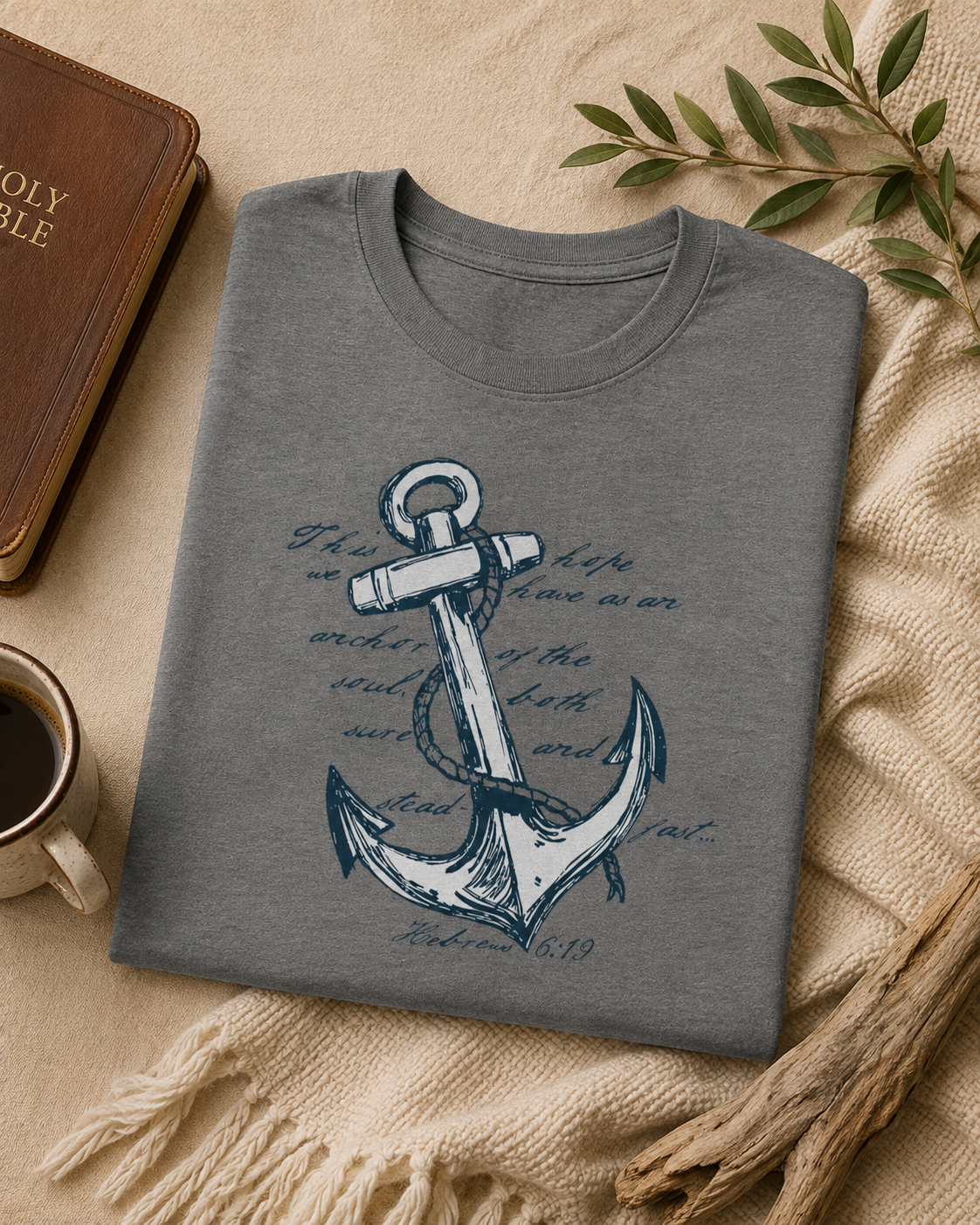 A folded heather gray t-shirt with a navy blue anchor graphic and Bible verse print, styled in a flat lay on a beige blanket with a book, coffee mug, and dried leaves.