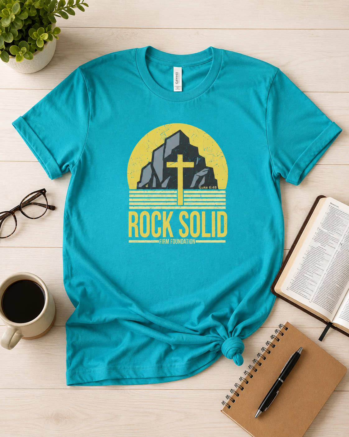 Teal Rock Solid Christian T-shirt flat lay styled with a Bible, coffee, eyeglasses, and journal