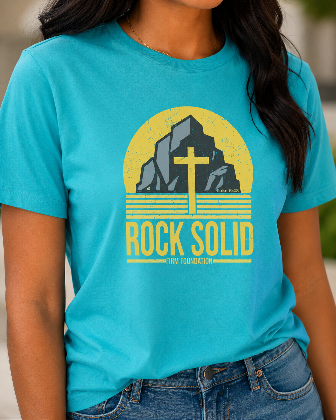 Close-up of a turquoise Rock Solid Christian T-shirt showing the Luke 6:48 cross and firm foundation design