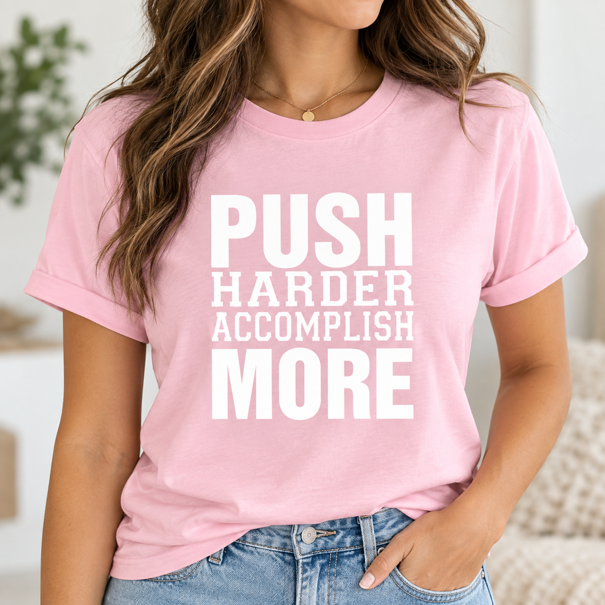 A woman wearing a light pink crewneck t-shirt with the motivational text 'PUSH HARDER ACCOMPLISH MORE' printed in bold white capital letters across the front, paired with blue jeans.