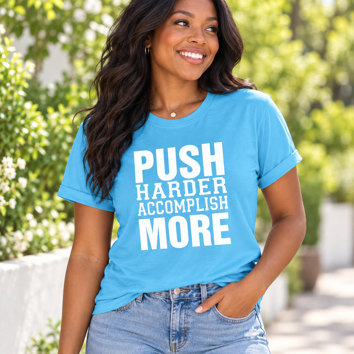 A woman wearing a bright blue graphic t-shirt with the text 'PUSH HARDER ACCOMPLISH MORE' in bold white letters, paired with light-wash blue jeans, set against a blurred outdoor background of green foliage.