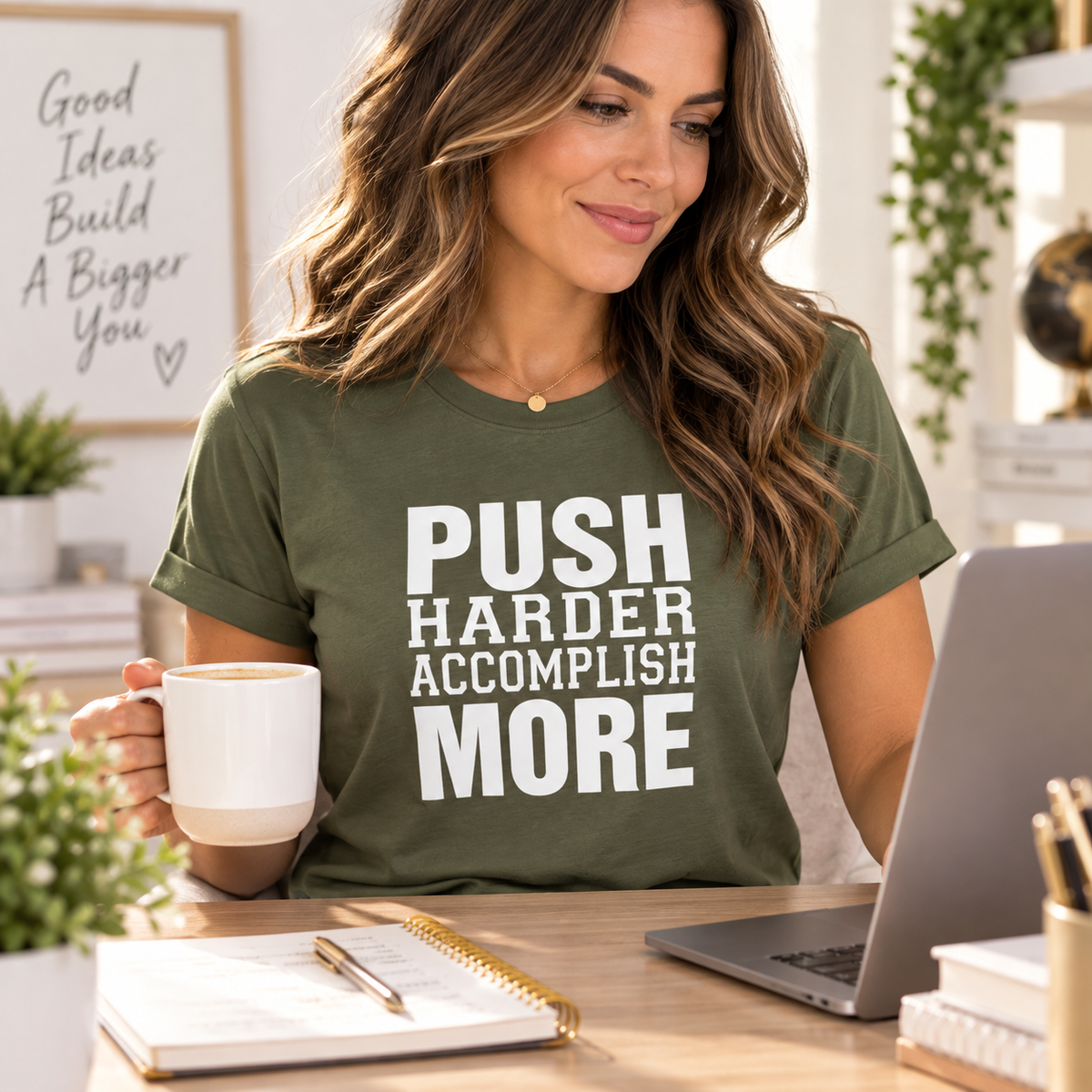 A woman sitting at a desk wearing an olive green graphic t-shirt with the text 'PUSH HARDER ACCOMPLISH MORE' in white. She is holding a white mug and looking down at a laptop in a bright, modern home office setting.