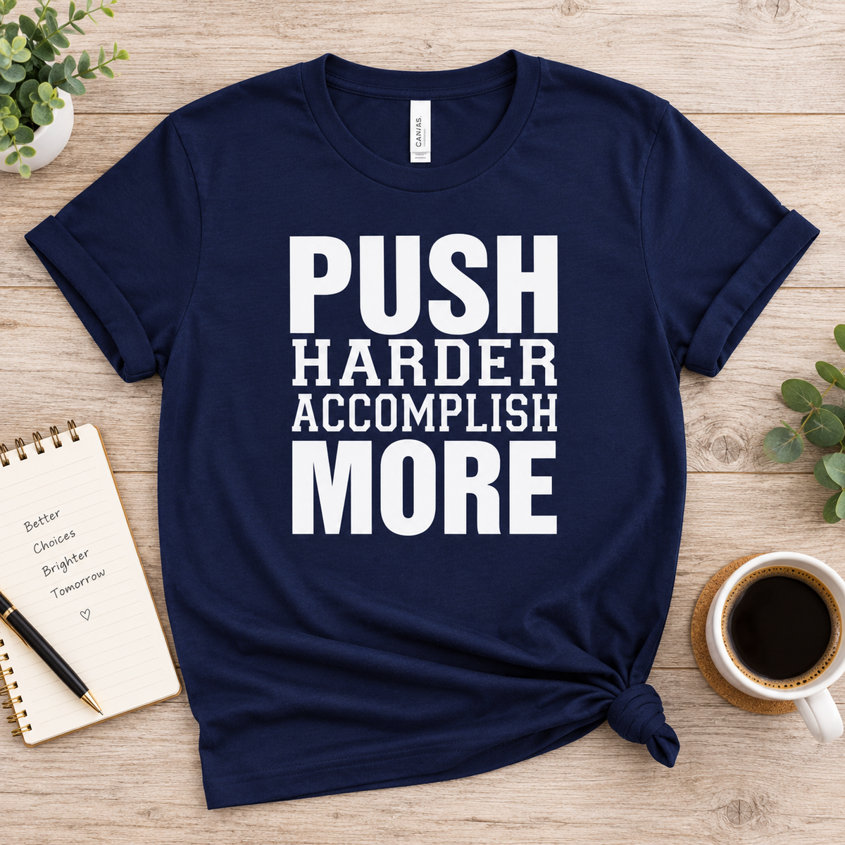 A navy blue graphic t-shirt with the text 'PUSH HARDER ACCOMPLISH MORE' printed in bold white capital letters, styled in a flat-lay on a light wood background with a coffee cup, notebook, and plants.