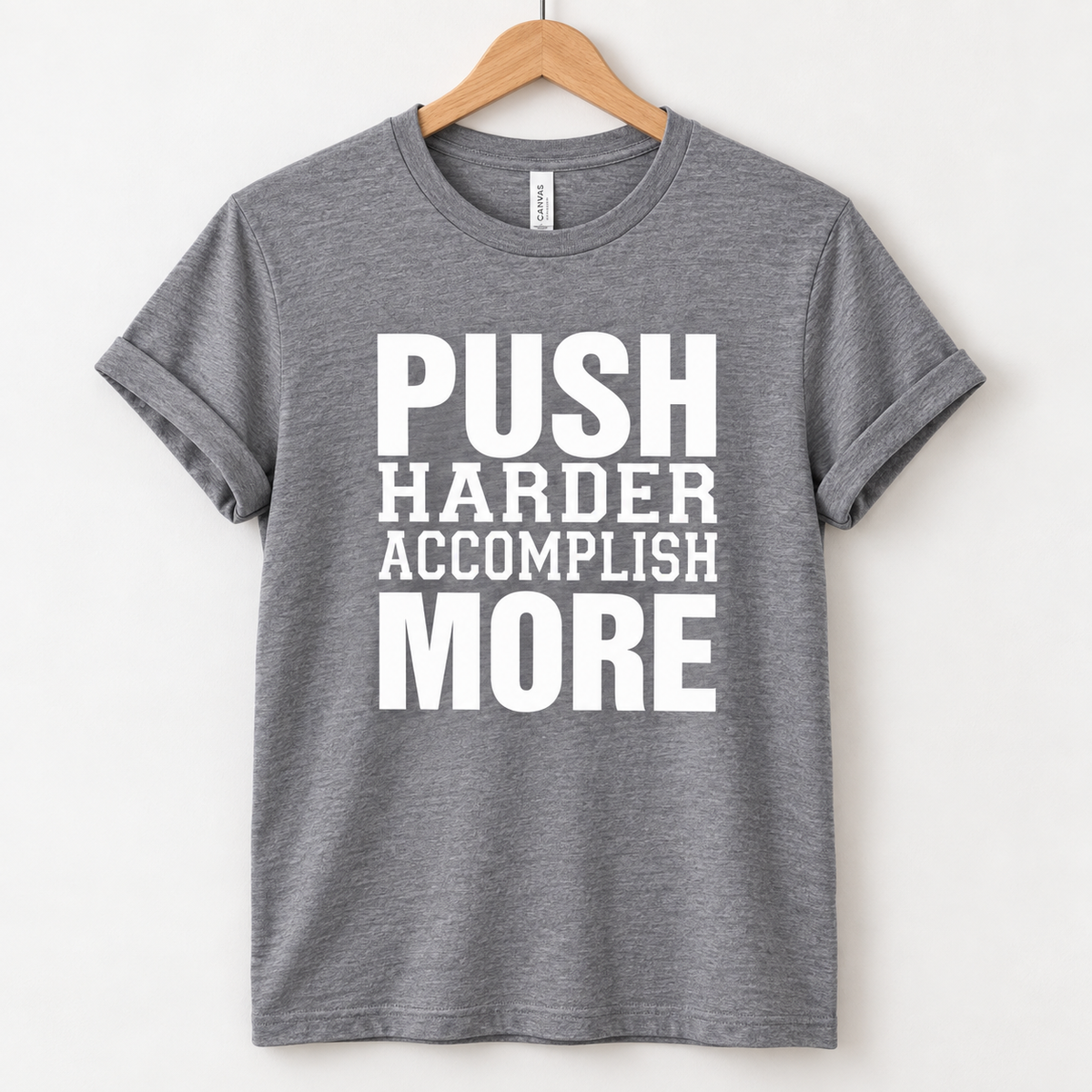 A heather gray short-sleeve t-shirt hanging on a wooden hanger against a light off-white wall. The shirt features a bold white graphic text print on the front that reads 'PUSH HARDER ACCOMPLISH MORE'.