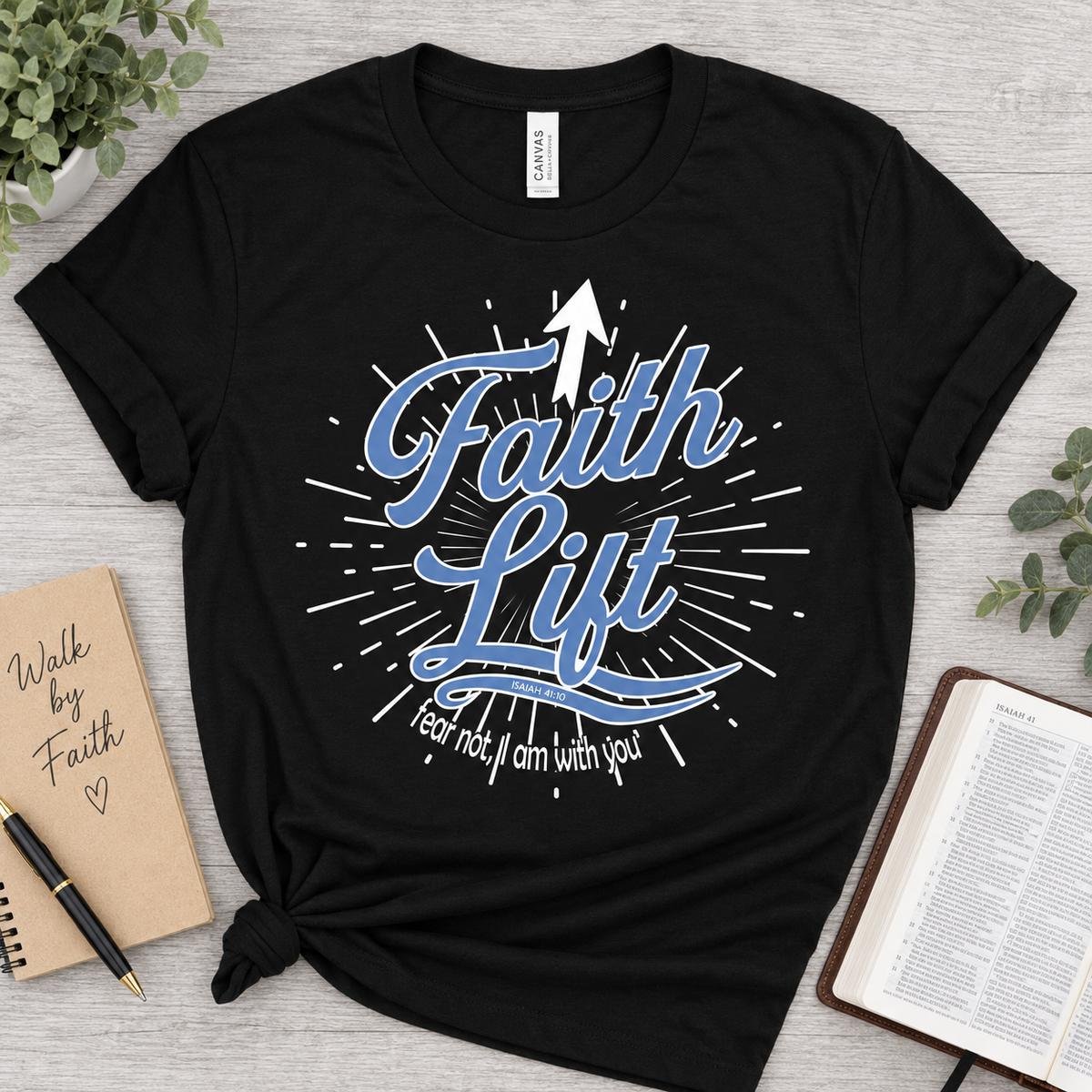 A black graphic t-shirt with a 'Faith Lift' design in blue and white, styled in a flat-lay arrangement with a notebook, pen, Bible, and greenery on a light gray wood background.