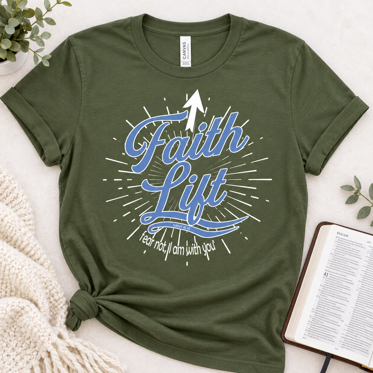 An olive green short-sleeve graphic T-shirt laid flat on a light fabric surface, styled with a knitted cream blanket, small greenery, and an open Bible. The shirt has a large blue and white 'Faith Lift' graphic with an upward arrow and the phrase 'Fear not, I am with you.