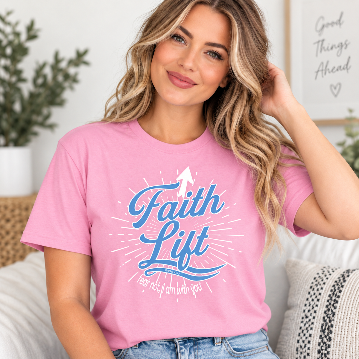 A woman wearing a pink graphic t-shirt with the words 'Faith Lift' in blue script and white rays, sitting on a couch in a bright living room.