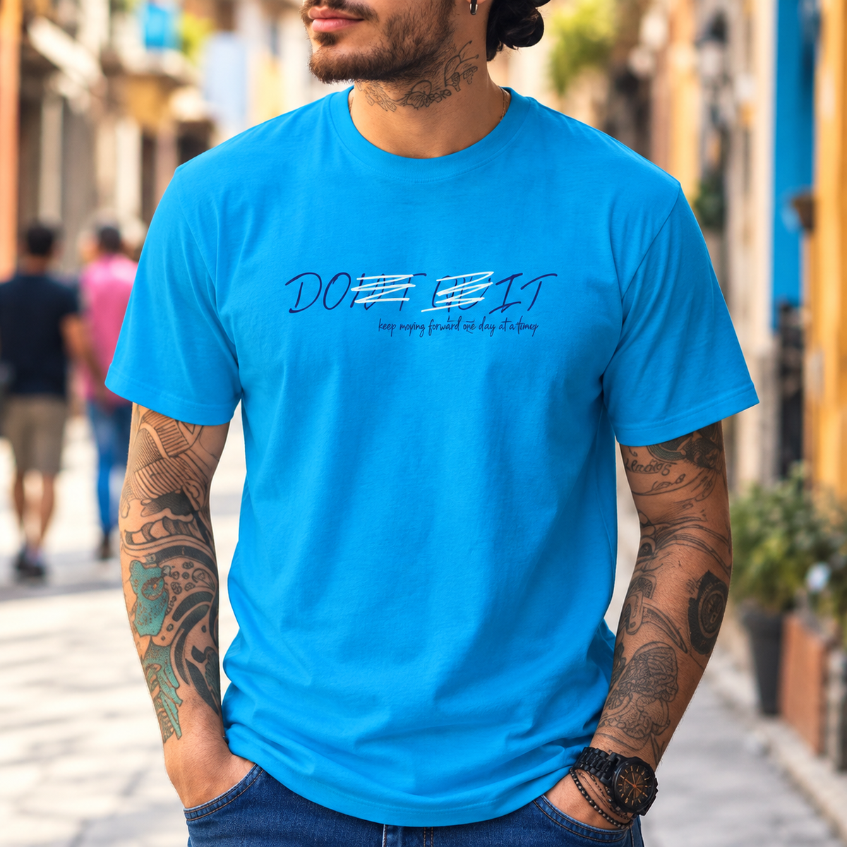 A man wearing a bright blue graphic t-shirt with the text 'DO IT' and 'keep pushing whenever life is hard, it's going to get easier.' He is standing in a blurred outdoor street setting.