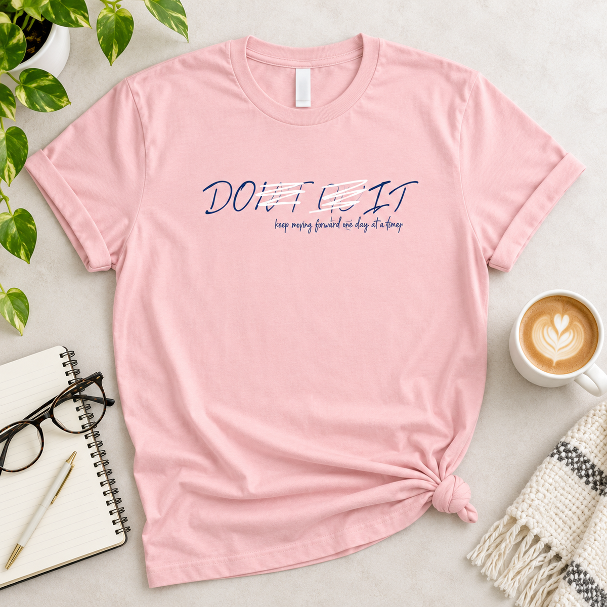 A light pink short-sleeve t-shirt with a blue graphic text print on the chest, styled in a flat-lay arrangement with a coffee cup, notebook, glasses, and green leaves on a light gray surface.
