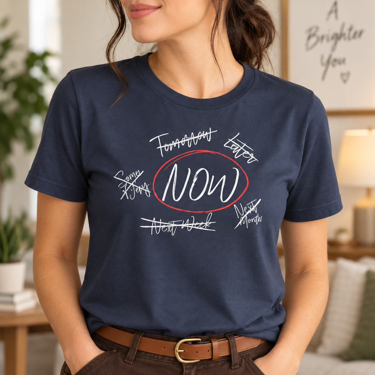 A woman wearing a navy blue graphic t-shirt with a motivational design that reads 'FUTURE DREAMER, NOW' in white and red script, set against a cozy, softly lit living room background.