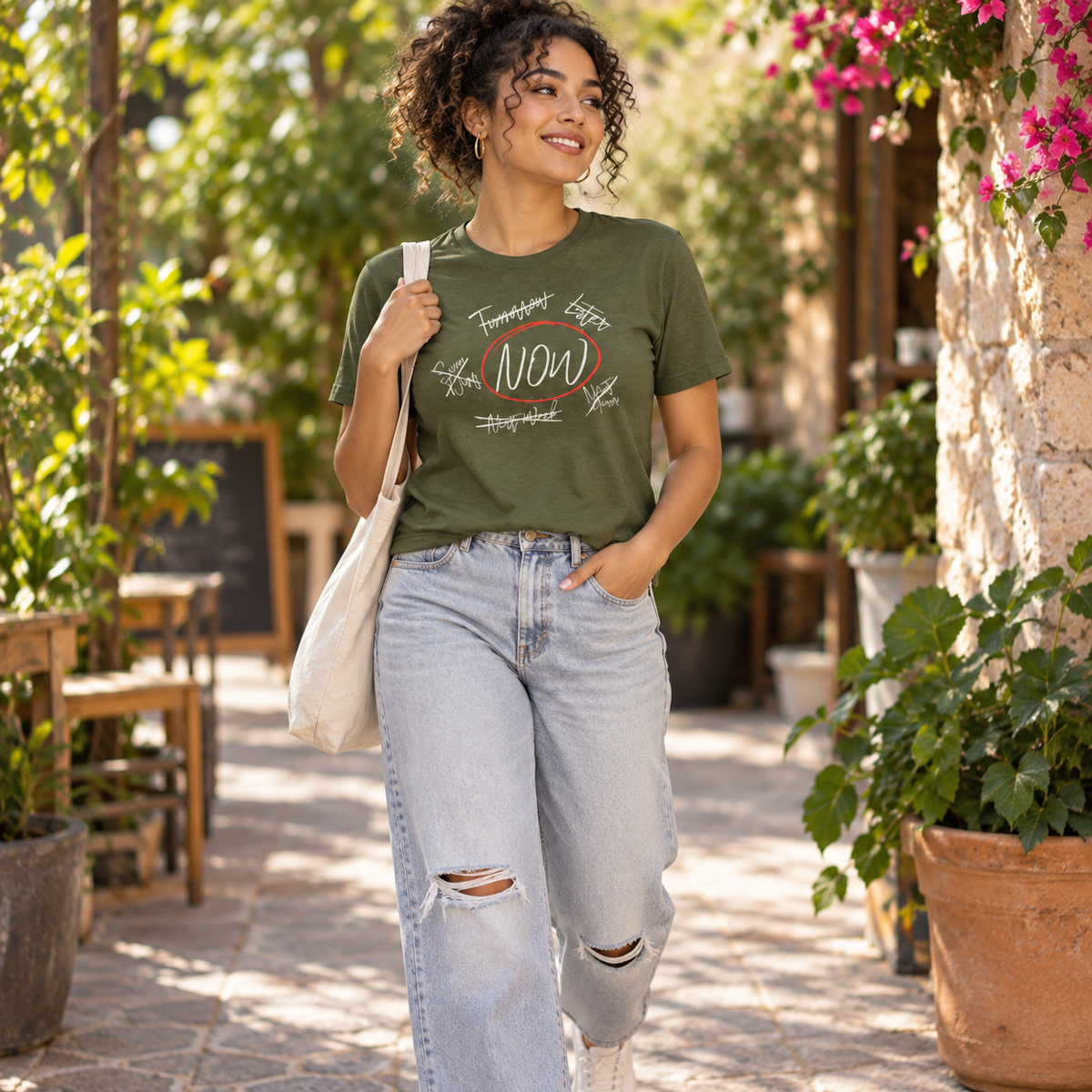 A woman stands outdoors in a garden courtyard wearing an olive green graphic t-shirt, light-wash ripped jeans, and carrying a cream canvas tote bag.