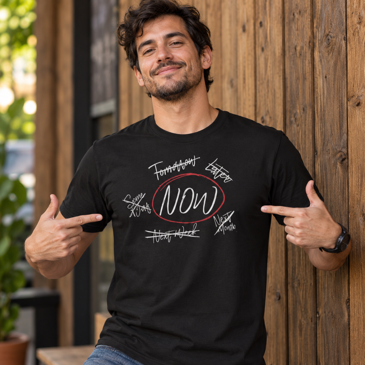 A man wearing a black graphic t-shirt with a 'NOW' motivational print, posing in front of a rustic wooden wall.