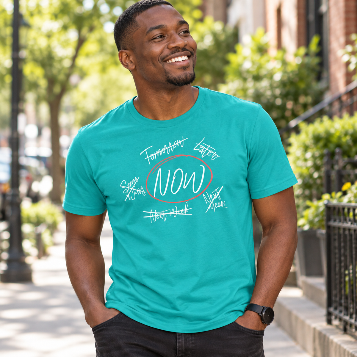 A man wearing a teal t-shirt with a white graphic print that reads 'FUTURE DATES, LATER NOW' circled in red, standing on a sunny city sidewalk.
