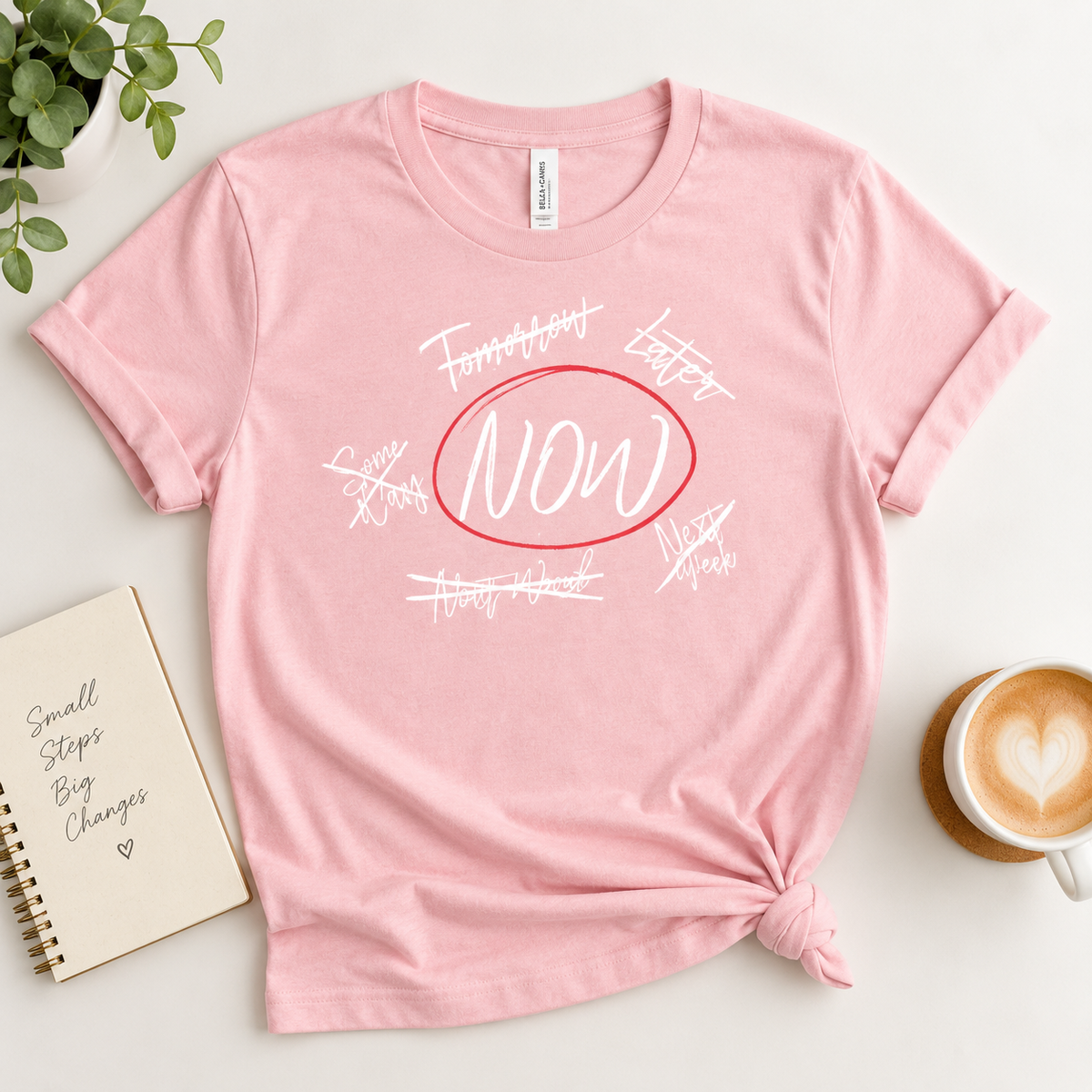 A pink short-sleeve graphic T-shirt laid flat on a light surface, styled with a plant, notebook, and coffee cup. The shirt has a red oval outline around the word “NOW” in white script, with additional white handwritten-style text around it.
