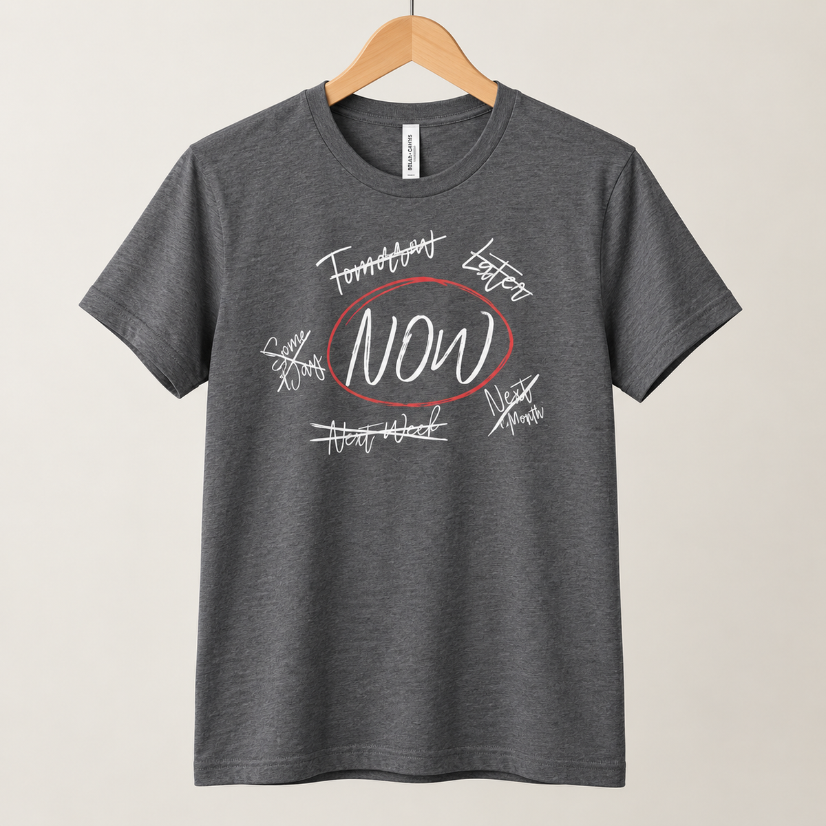 A dark heather gray t-shirt hanging on a wooden hanger against a light beige background. The shirt features a graphic print on the front with the word 'NOW' in white script inside a red oval, surrounded by white cursive text signatures.
