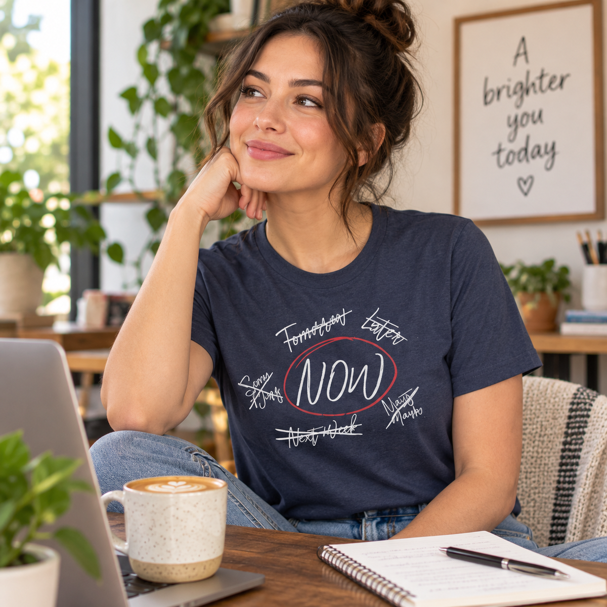 A woman sitting at a desk in a bright, plant-filled room wearing a navy blue graphic t-shirt with white handwritten-style text and a red circle around the word 'NOW'.