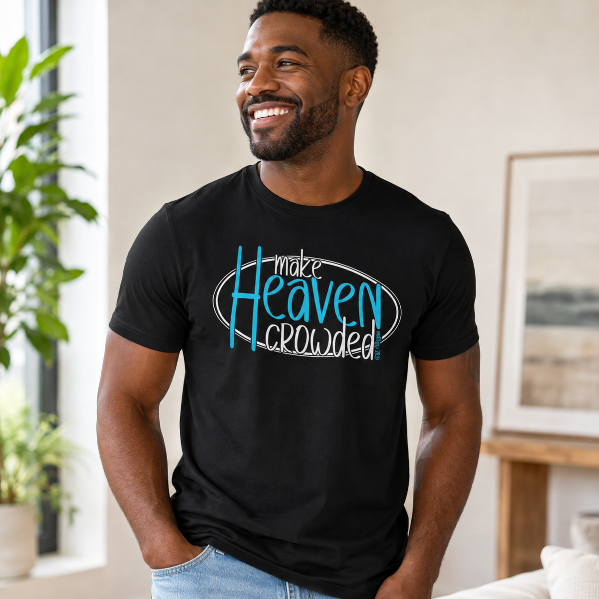 A smiling man wearing a black graphic t-shirt with the text 'make Heaven crowded' in blue and white, standing in a brightly lit room with a plant and framed art in the background.