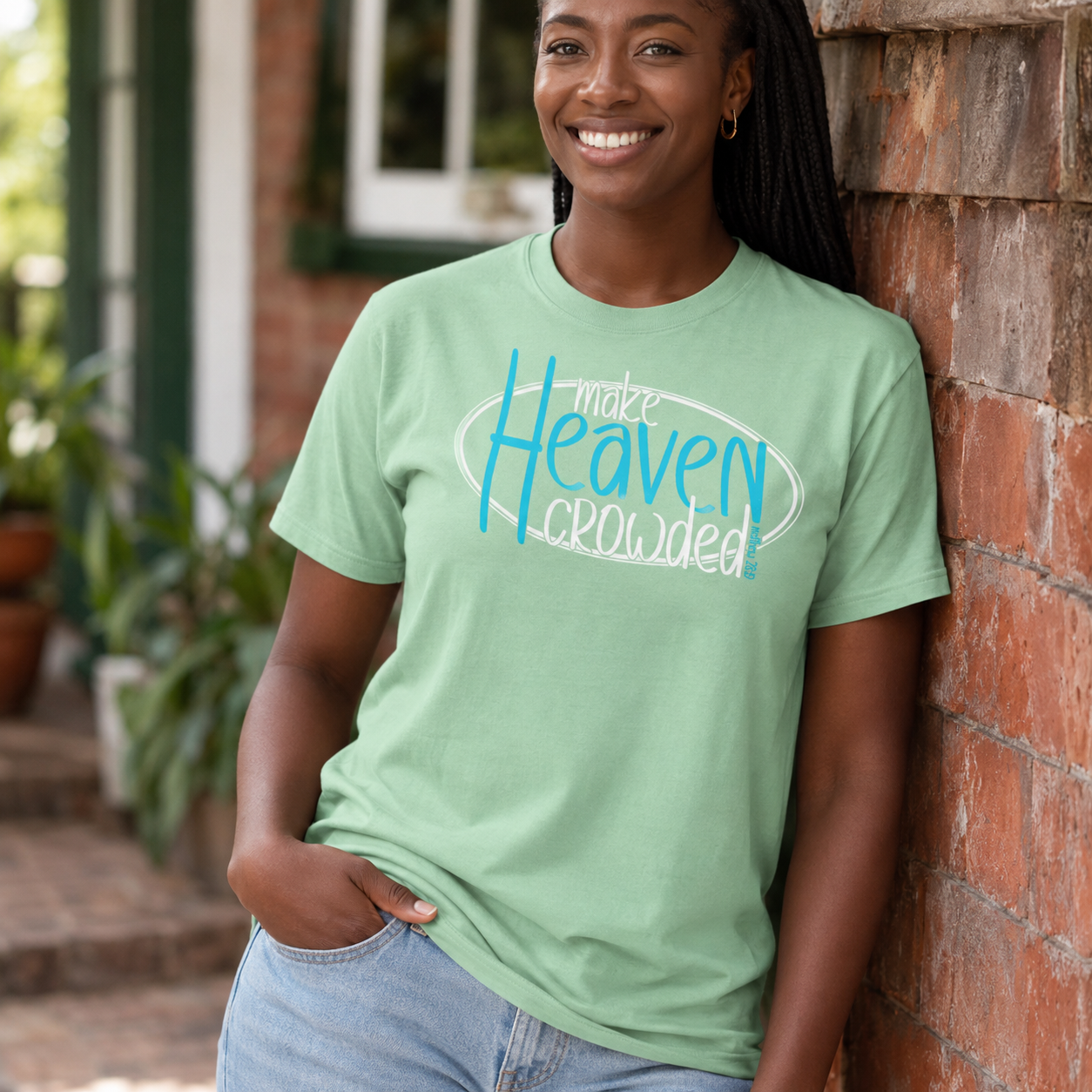A woman wearing a sage green graphic t-shirt with the text 'make Heaven crowded' in blue and white, leaning against a brick wall outdoors.