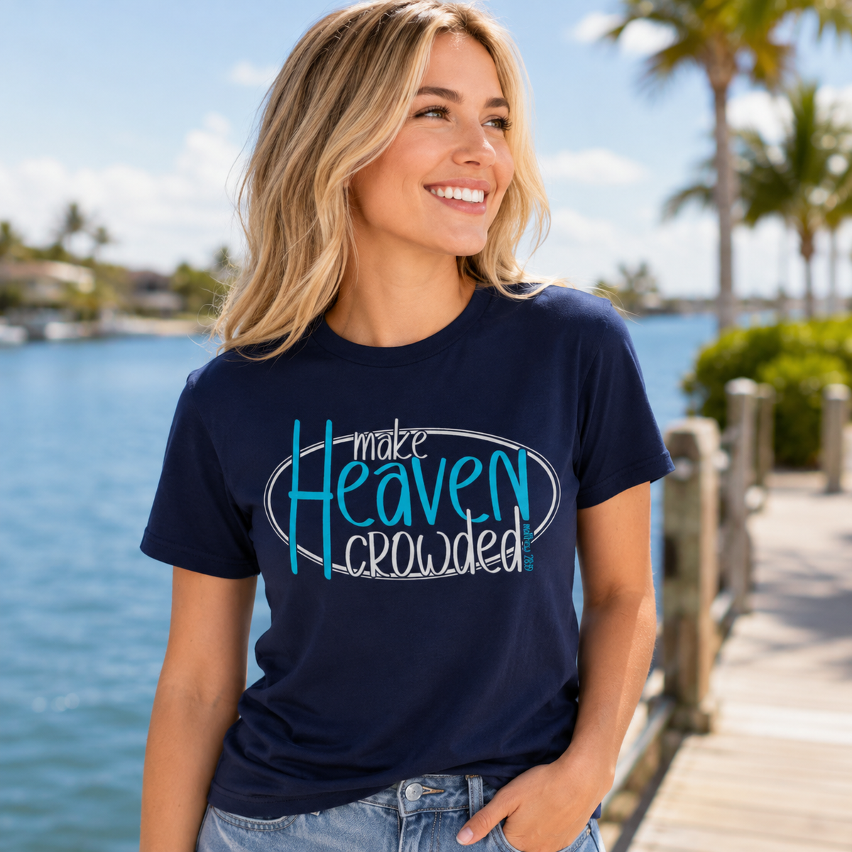 A woman wearing a navy blue graphic t-shirt with the text 'make Heaven crowded!' in blue and white, standing on a wooden dock with a tropical waterfront background.