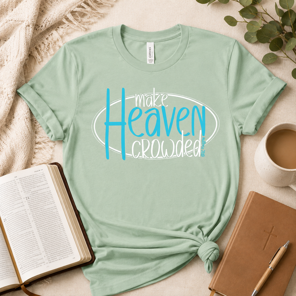 A mint green graphic t-shirt with the text 'make Heaven crowded!' in blue and white, styled in a flat-lay arrangement with a Bible, a coffee mug, a notebook, and a knotted fabric detail at the hem.