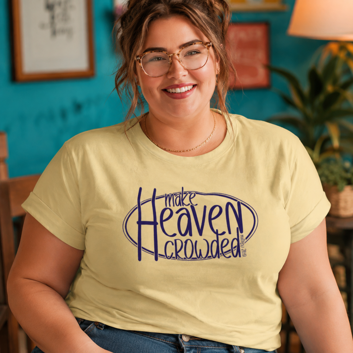 A woman wearing a pale yellow graphic t-shirt with the text 'make Heaven crowded' in dark blue, posed in a cozy indoor setting with a teal wall and warm lighting.