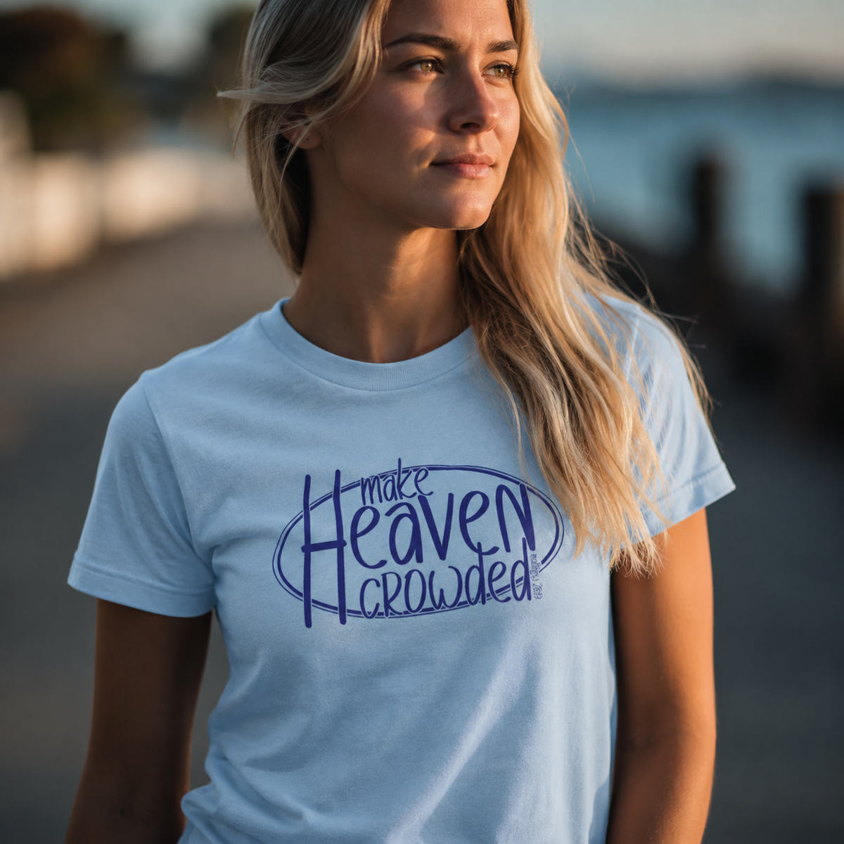 A woman wearing a light blue crewneck t-shirt with a dark blue graphic that reads 'make Heaven crowded' in a stylized font, photographed outdoors with a blurred background.
