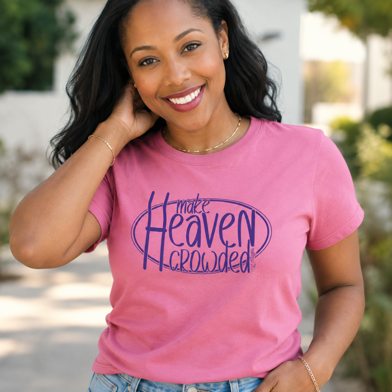 A smiling woman wearing a pink short-sleeve graphic T-shirt with the phrase 'make Heaven crowded' printed in dark purple script and oval outline, photographed outdoors with a softly blurred background.