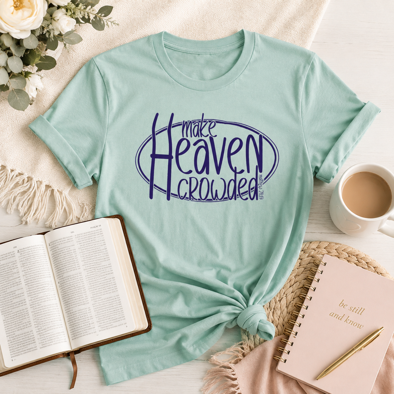 A mint green graphic t-shirt with the text 'make Heaven crowded' in dark blue, styled in a cozy flat-lay arrangement with a coffee mug, open Bible, notebook, and flowers.