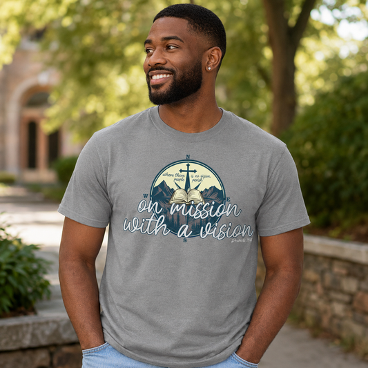 A man wearing a heather gray graphic t-shirt with a blue and white 'on mission with a vision' design, standing outdoors in a park-like setting with blurred green trees and stone walls.