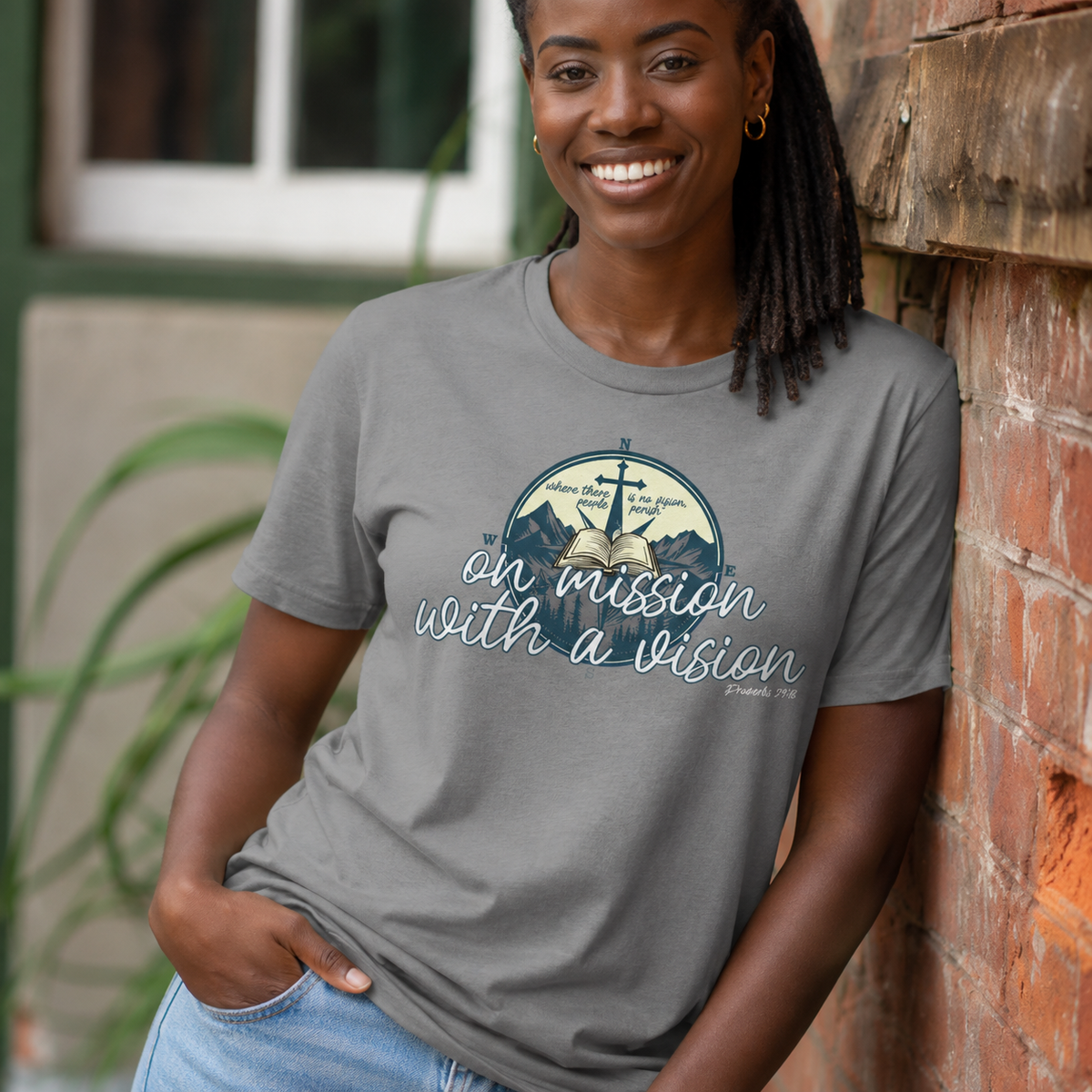 A smiling woman with dreadlocks leans against a brick wall wearing a heather gray graphic t-shirt. The shirt features a circular graphic with mountains, an anchor, and the text 'on mission with a vision' in white script.
