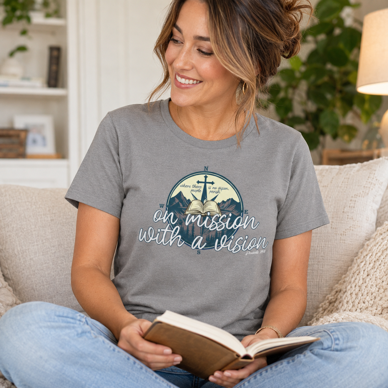 A woman sitting on a beige couch in a cozy living room setting, wearing a heather gray graphic t-shirt with a circular mountain and compass design that reads 'on mission with a vision'. She is holding an open book.