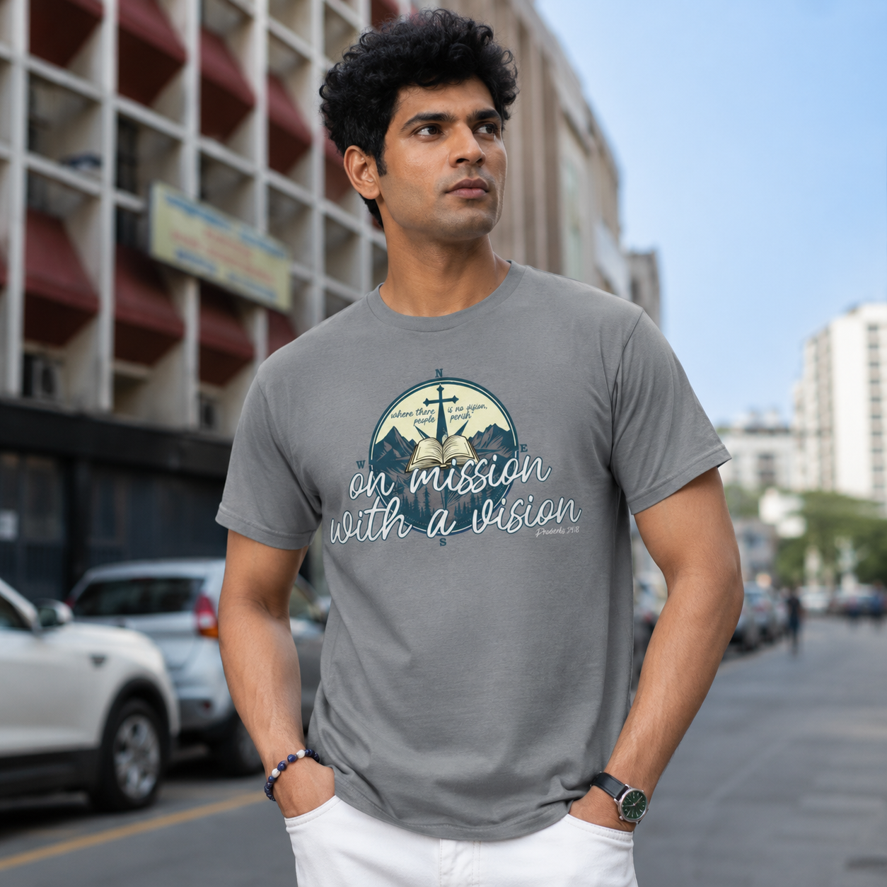 A man wearing a gray graphic t-shirt with a nautical anchor and ship design and the text 'an mission with a vision' stands on a city street.