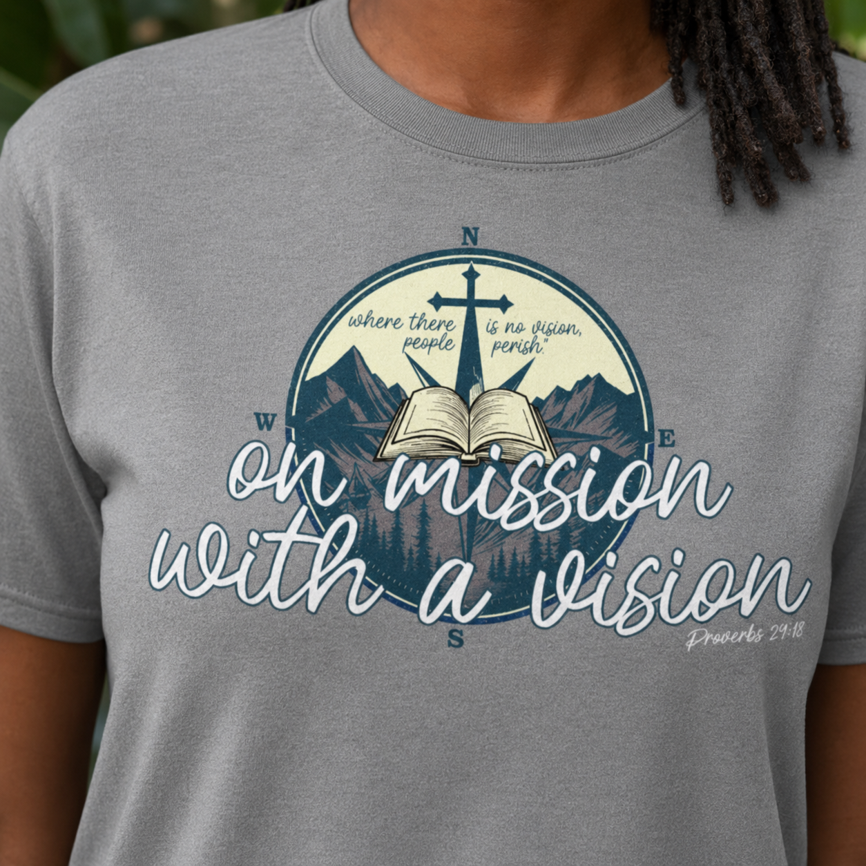 A close-up of a person wearing a heather gray t-shirt with a graphic print. The graphic features a compass with a cross, an open Bible, mountains, and pine trees, with the text 'where there are people, there is no vision, without God, we are lost' and 'on mission with a vision' in white script.